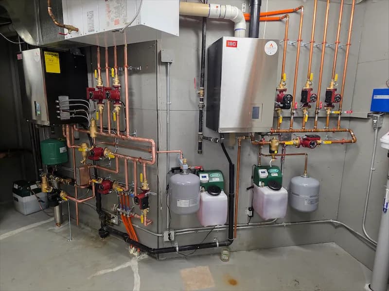 Wall-mounted condensing boiler beside a copper manifold with red-handled zone valves, circulator pumps and glycol feed bottles in a residential mechanical room