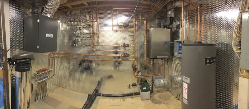 Wide view of a finished mechanical room with overhead copper heating lines, a control panel and a hot water tank